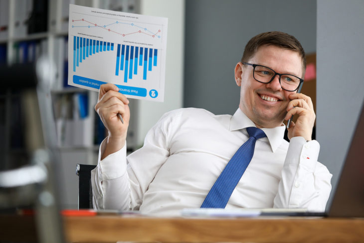 Smiling man talking on the phone while holding a document with charts.